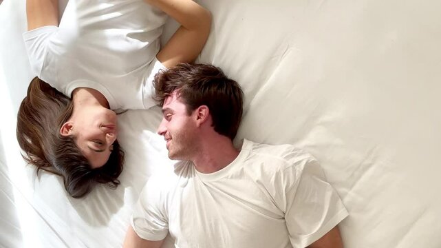 A millennial couple lying in bed with white T-shirts and sheets, talking happily and calmly about relationship issues, future plans, and mutual trust during a serene morning. People in bed at morning