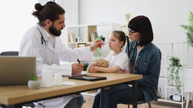 Caucasian male doctor measures temperature of little sick Caucasian girl sitting with mother. Doctor uses thermometer for accurate reading, while mother provides comfort to daughter holding teddy bear