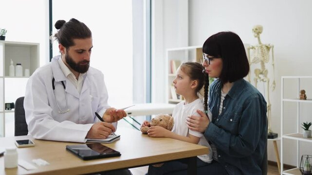 Caucasian male doctor in white coat measures temperature of little sick Caucasian girl sitting on mother's lap in clinic. Doctor uses digital thermometer during medical examination.
