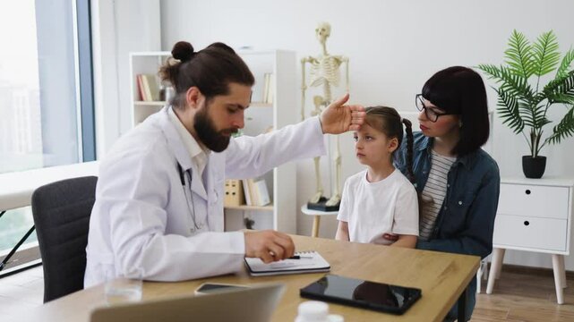 Caucasian male doctor checks temperature of sick young girl while mother looks on in medical office. Doctor is examining girl's forehead, showing concern and care.