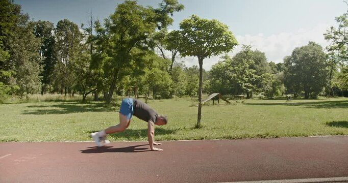 A muscular man performs jumping jacks and burpees near a running track in a park.