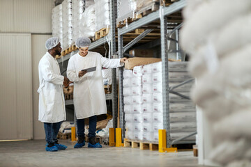 Full length of arab warehouseman with tablet preparing sacks with flour for black worker in food factory.