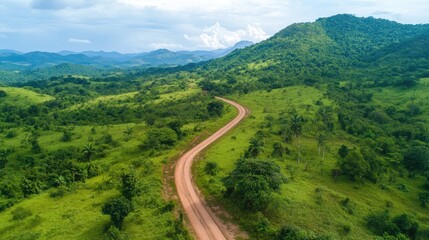 Fototapeta premium Aerial view of a winding rural road meandering through vibrant green fields and rolling hills, framed by a backdrop of distant mountains under a cloudy sky.