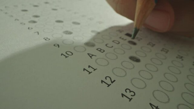 Macro close up of Unrecognizable hands filling up answer sheet with a pencil in classroom. Student shading circles correct answers in standard multiple choice exam test paper. Educational questions.