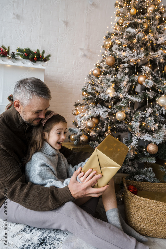 Wall mural Father and daughter excitedly open a gift, their expressions filled with wonder and joy. The Christmas tree adds to the festive atmosphere