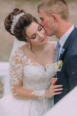 A bride and groom are kissing each other's lips. The bride is wearing a white dress and a tiara....