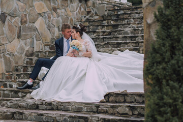 A bride and groom are sitting on a stone staircase. The bride is wearing a white dress and the groom is wearing a suit. They are holding hands and looking at each other