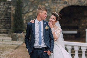 A bride and groom are standing together in front of a stone building. The bride is wearing a white dress and a veil, while the groom is wearing a suit and tie. They are both smiling