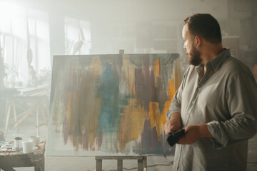 Male artist thoughtfully looking at a freshly completed painting in his studio.