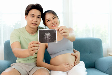 Fototapeta premium Happy family at home, Asian couple show ultrasound baby picture while sit on sofa at living room together. Beautiful pregnant wife with big belly and her husband show her fetal photo. pregnancy mother