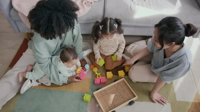 Above shot of young Asian woman passing yellow cube to her daughter building tower while both sitting on the floor in front of African American mother with baby son