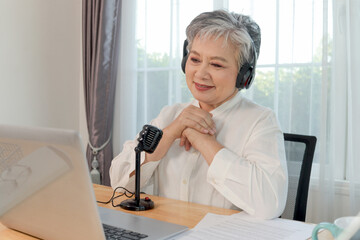 Happy Asian elderly woman influencer with headphones and laptop speaks in microphone for recording...