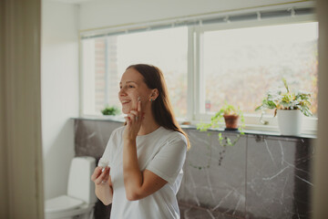 Happy Woman Applying Face Cream Moisturizing And Caring For Skin Smiling To Her Reflection In Mirror Standing In Modern Bathroom At Home. Facial Skincare And Pampering. Selective Focus