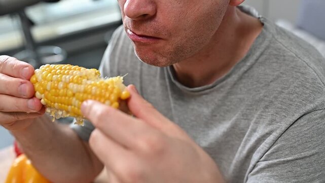 Close-up of Caucasian man eating yellow juicy corn cob eats with appetite in kitchen enjoy vegetarian dishes, organic healthy food