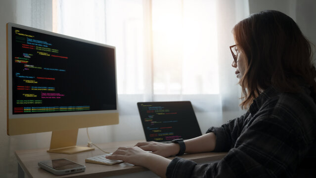A woman is sitting at a desk with a computer monitor and laptop in front of her. She is typing on the keyboard and looking at the computer screen. Scene is focused and determined