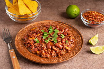 Mexican homemade savory spicy chili con carne dish stewed with beans, ground beef, tomatoes and chilli pepper seasoning served in plate on table with nachos tortilla chips, lime slices and fork