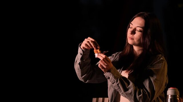 Cheerful hungry lady is dipping small piece of hamburger into disposable sauce package and chew it with delight. Slow motion view of Caucasian woman eating junk food at night time in the street