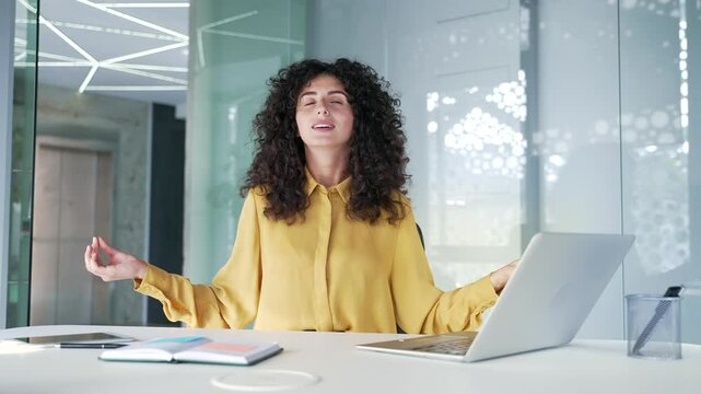 Young adult businesswoman is meditating with her eyes closed sitting at workplace in office. Female employee took break from work to relieve stress. Brunette woman practices yoga, feels peace of mind