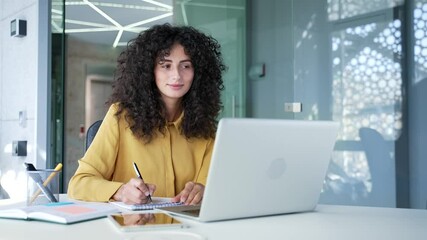 Young adult female employee watching video call training notes in notebook looking at laptop computer screen sitting in business office. Businesswoman is studying online, listening remotely to course
