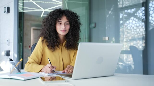 Young adult female employee watching video call training notes in notebook looking at laptop computer screen sitting in business office. Businesswoman is studying online, listening remotely to course