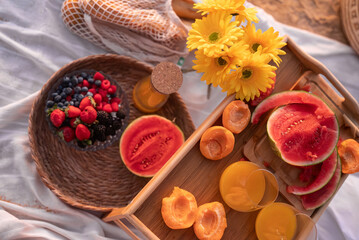 A vibrant summer picnic setup featuring fresh watermelon, assorted berries, apricots, and juice. Bright yellow flowers add a cheerful touch to this inviting outdoor scene.