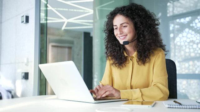Young adult woman in wireless headset customer service representative operator talking on video call on laptop sitting in business office. Agent of the call center advises client, answering questions