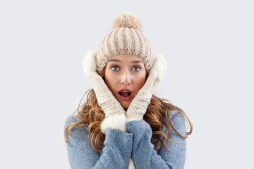 Amazed woman embracing herself in winter Christmas time. Wearing a cozy blue sweater, pom-pom beanie hat with ear warmers, scarf, and gloves. She smiling at the camera, isolated on a gray background