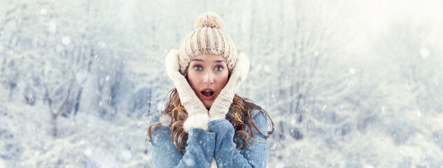 Amazed woman enjoying winter snowfall outdoors in nature. She marvels at the falling snow during a white Christmas. Wearing a cozy blue sweater, pom-pom beanie hat with ear warmers, scarf, and gloves