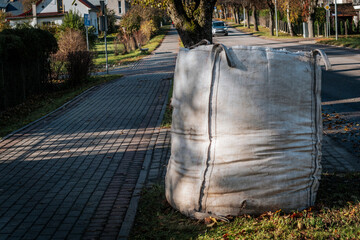 A close-up of a large, white cloth bag placed on a paved area by the roadside. The bag appears to...