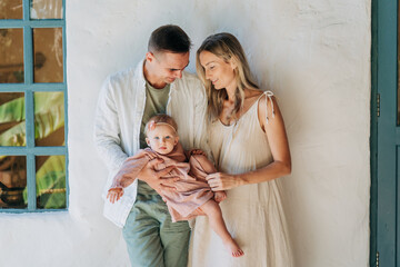 Mother and father smile and hold their little daughter standing against the wall.