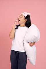 A beautiful Asian woman, tired from a day of sleep deprivation in white pajamas and an eye mask, is yawning with her hand covering her mouth, standing on a pink background.