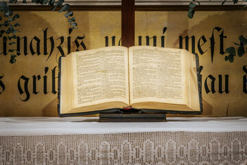 A church altar featuring an open Bible, a wooden crucifix, and floral arrangements on either side. The background includes a religious painting and decorative script