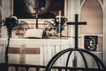 An iron cross stands prominently in the foreground with a blurred view of an open Bible and church altar in the background. The scene reflects a solemn atmosphere of faith, prayer, and worship inside 