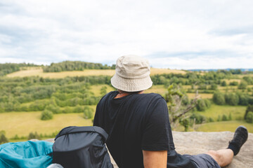 Person wearing a beige bucket hat and a black shirt sits on a rock, gazing at the vast green landscape and rolling hills of the Beskydy Mountains on an overcast day