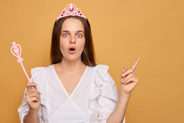 Young woman in white dress and pink tiara points excitedly while holding a magic wand, on a bright...