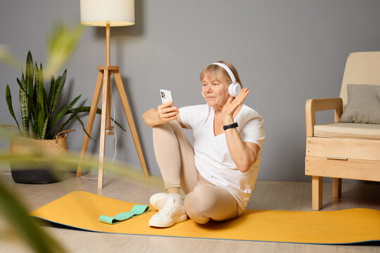Older woman sitting on a yoga mat at home, using a smartphone and wearing headphones while enjoying a virtual fitness session in a cozy room with plants