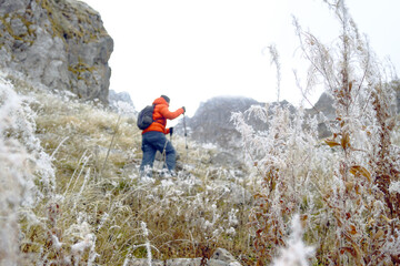 Fototapeta premium A tourist walks in the mountains during the cold season: a landscape with a man in a bright jacket walking up using hiking poles and frost-covered plants (Prokletije National Park, Montenegro).