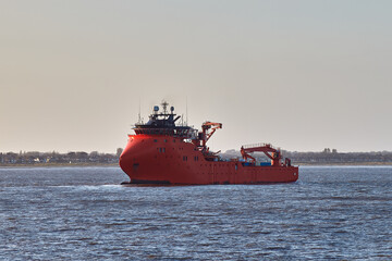 Red offshore support vessel sailing in port on a bright day.