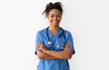 Portrait of positive female African American doctor or nurse posing with crossed arms and smiling looking at camera isolated over white studio background wall, lady wearing blue coat and stethoscope