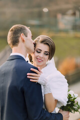 A bride and groom are hugging each other, with the bride wearing a white fur stole. The man is kissing the woman's neck
