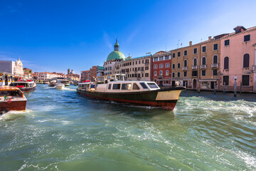 A large public transportation vaporetto near Ferrovia stop, moves along the Grand Canal in Venice. Other smaller boats are visible, along with the historic architecture of the city. Vibrant blue sky.