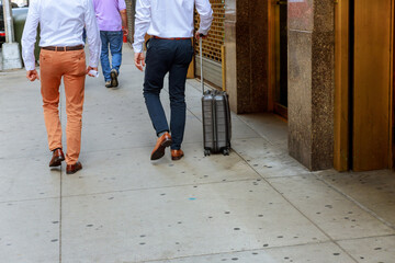 Fototapeta premium Two men in dress shirts, slacks stroll along busy city sidewalk, one pulling carry on suitcase, indicating travel professional meeting nearby.