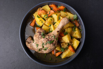 Duck leg with broth, stewed with potatoes and carrots, sprinkled with parsley and sumac. Simple home hearty cooking. Deep gray plate on a black background. Selective focus, close-up.