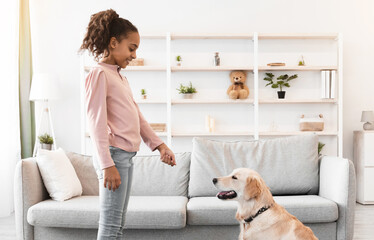 Pet Care And Love Concept. Side view portrait of smiling African American girl playing with her dog, feeding golden retriever with treats, training at home in living room, modern house interior