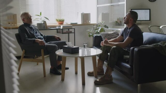 Mature male therapist listening intently to soldier in military uniform, sitting in front of him on couch, reliving his traumatic experience, while talking emotionally about past events at therapy