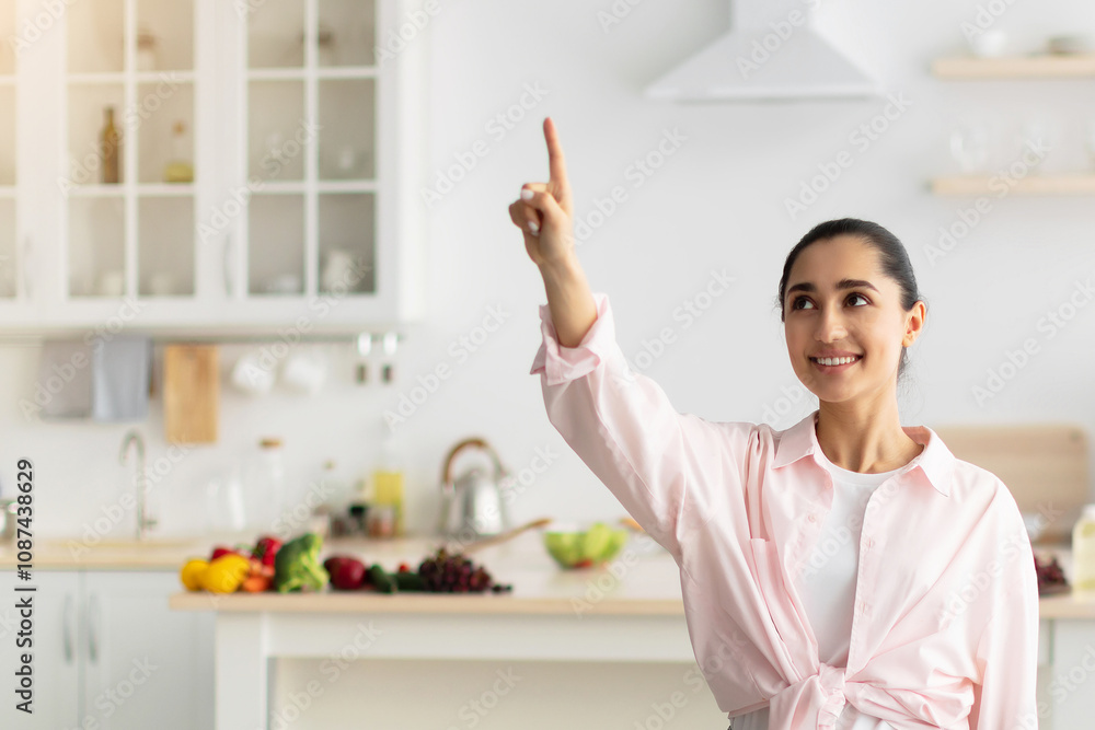 Aufkleber Modern Futuristic House Control System, IoT Technologies Concept. Portrait Of Smiling Millennial Lady Pointing Finger Up In The Air Pushing Button On Invisible Screen Standing In Kitchen Interior