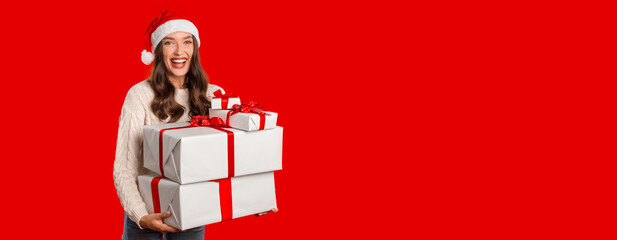 Joyful young lady in Santa hat holding two Xmas present boxes on red studio backdrop, greeting expressing positivity and merry holiday vibes. Christmas gifts delivery and celebration, copy space