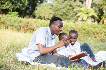 A father enjoys quality time with his young sons, reading a storybook together during a relaxing picnic in picturesque park surroundings.