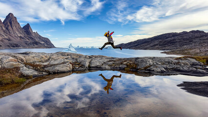 Young man jumping across rocks in Bear Islands, Scoresby Sund, Greenland. There is a perfect mirror reflection in the calm fjord waters, and mountains and icebergs can be seen in the distance. © Rixie