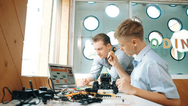 Caucasian teacher help student fixing car model and giving advise for programing system or coding prompt while highschool boy listen and use equipment to repair robot at STEM classroom. Edification.